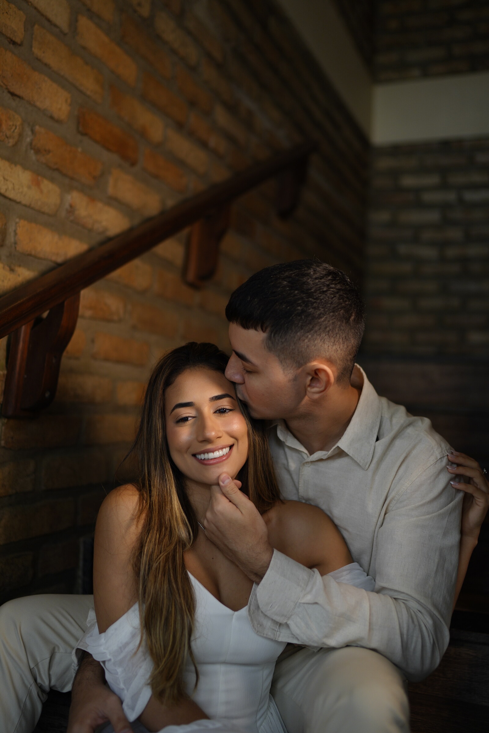 Danielle e Felipe sentados numa escada, junto a uma parede de tijolos; ele beija seu rosto