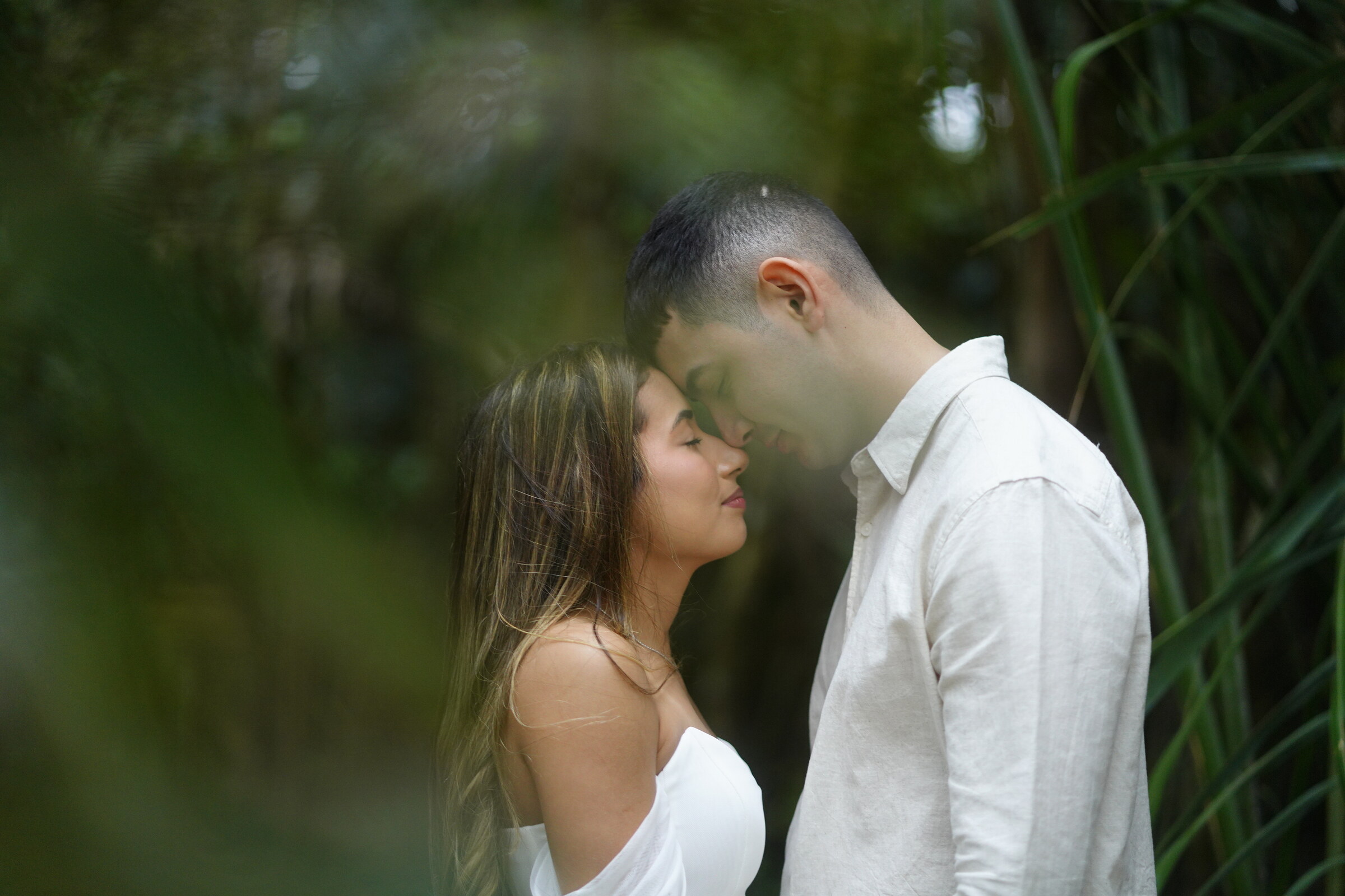 Danielle e Felipe de testa colada, entre as folhagens de um jardim de mata fechada